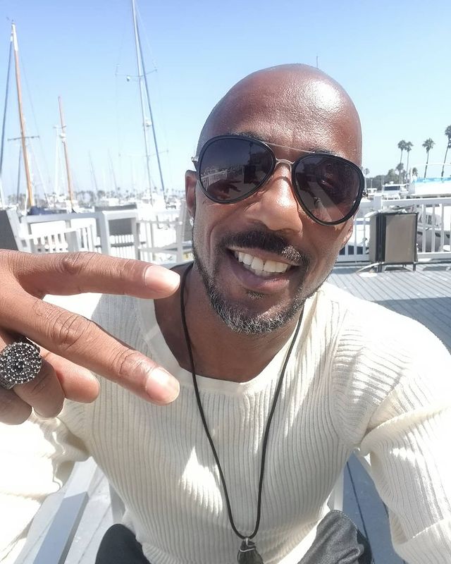 He is wearing white sweat-shirt with black chain and black goggles.